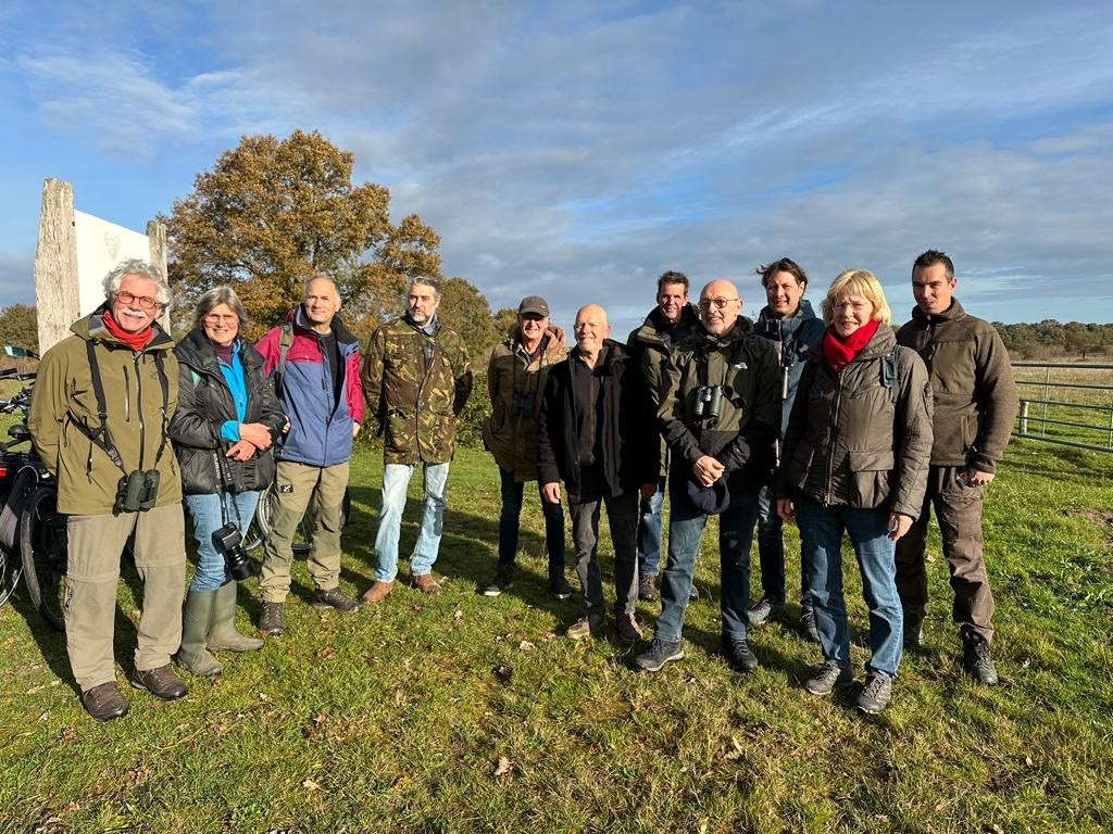 Groep mensen in outdoor-kleding poseert in een grasveld met een boom en blauwe lucht op de achtergrond.