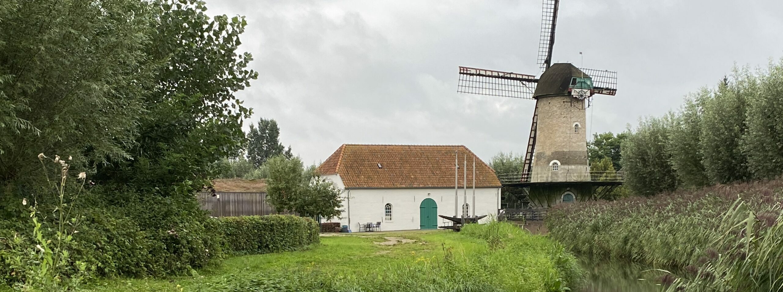 Windmolen en huisje naast een kanaal, omringd door bomen en begroeiing onder een bewolkte hemel.