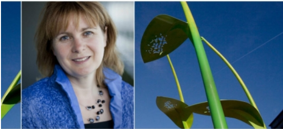 Portret van een vrouw in blauw, naast groene sculptuur tegen een heldere lucht.