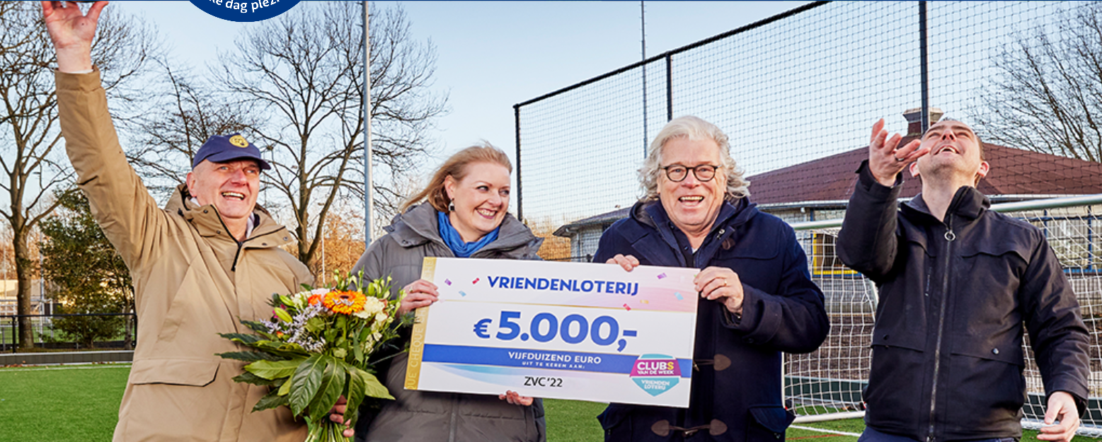 Vier mensen vieren een winstsituatie op een sportveld met een cheque van €5.000 en bloemen.