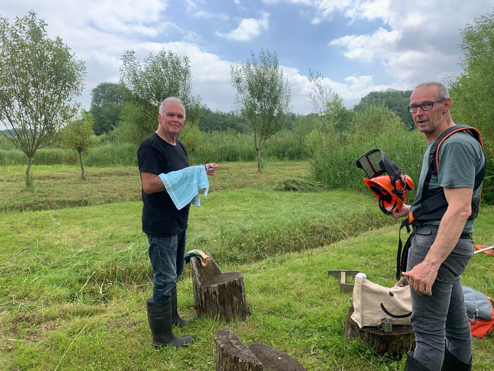 Twee mannen op grasveld, één met handdoek, andere met helm, omringd door bomen.