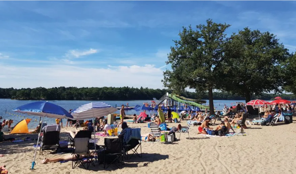 Strand aan meer met veel mensen, parasols en een speeltuin, onder een heldere blauwe hemel.