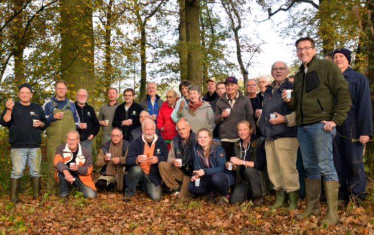 Een groep mensen in een herfstbos, met laarzen en bekers, poseren voor een foto.