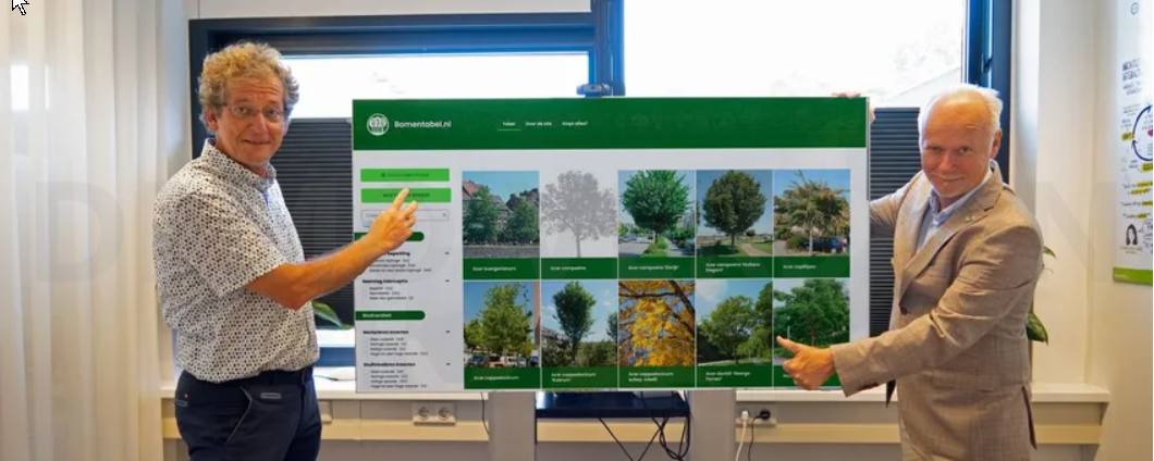 Twee mannen presenteren een poster met boominformatie en afbeeldingen in een verlichte kamer.