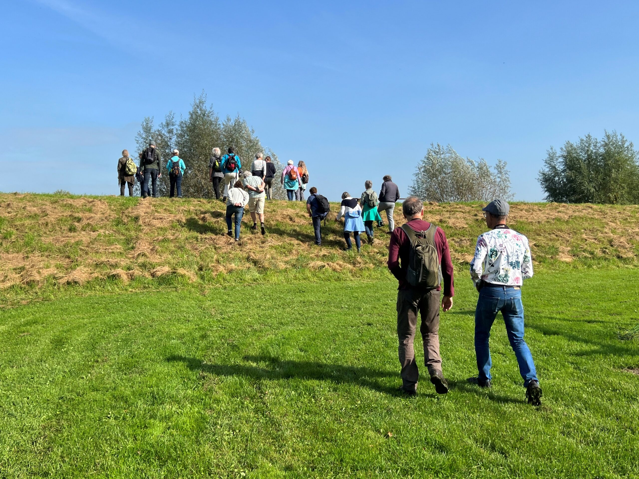 Groep mensen wandelt een grasheuvel op onder een heldere blauwe lucht.