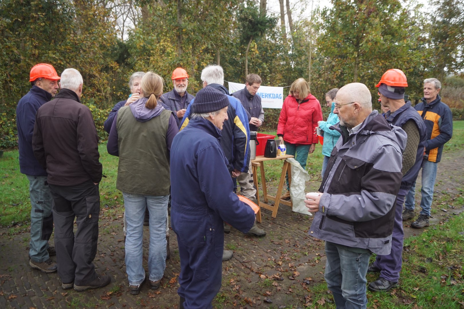 Groep mensen met oranje helmen heeft een pauze tijdens een werkdag in een groene buitenomgeving.