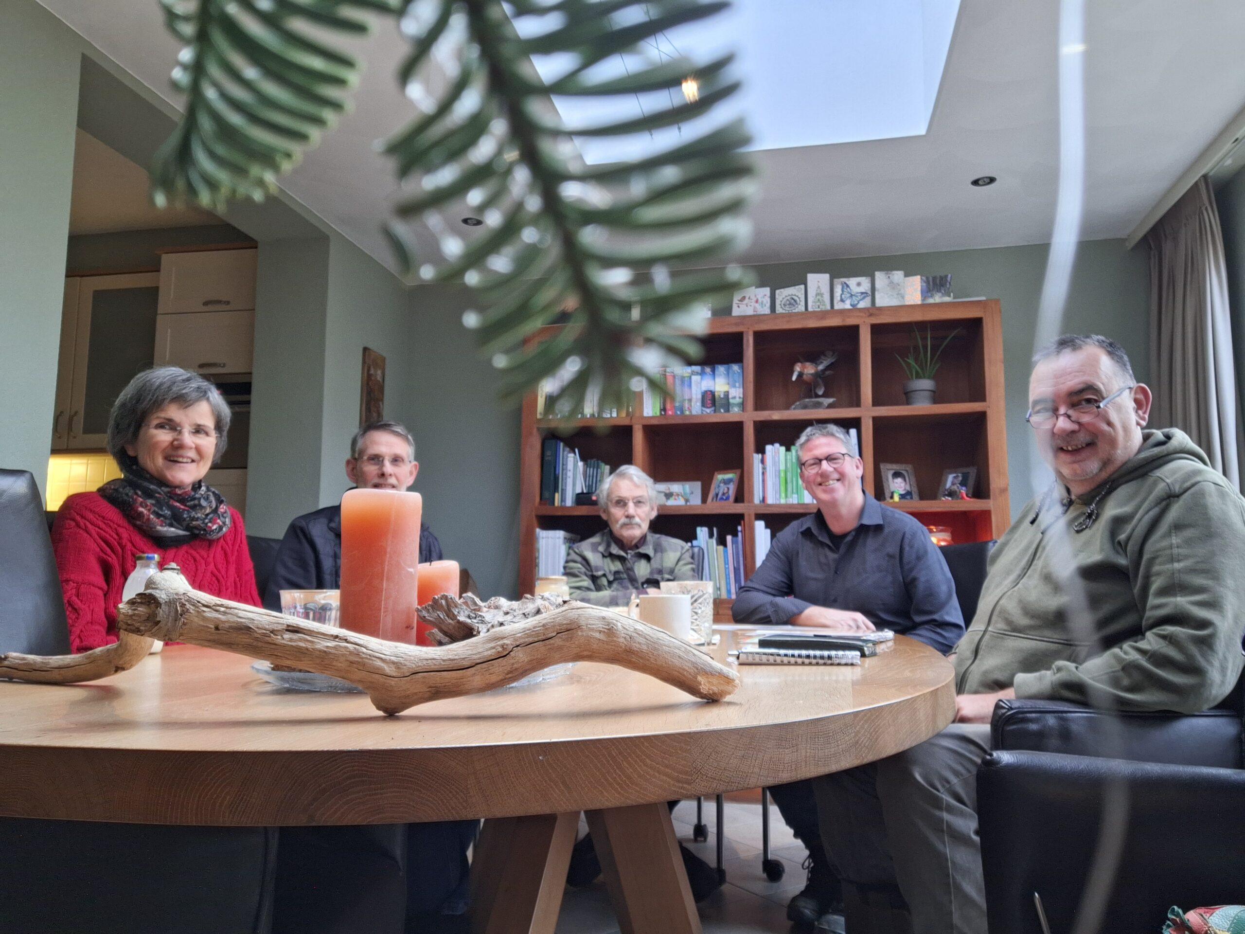Groep van vijf mensen zit lachend aan een ronde tafel met kaarsen en boekenplank op de achtergrond.