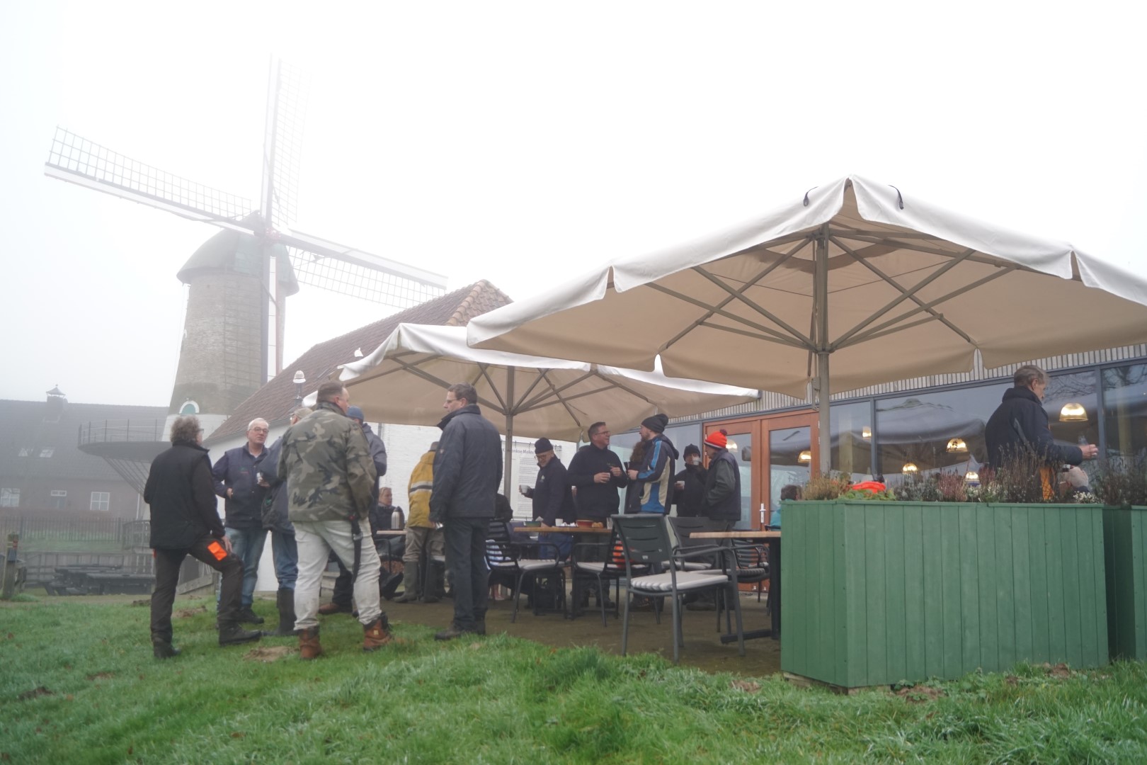 Mensen verzamelen zich onder parasols bij een molen; het is mistig en er hangt een gezellige sfeer.