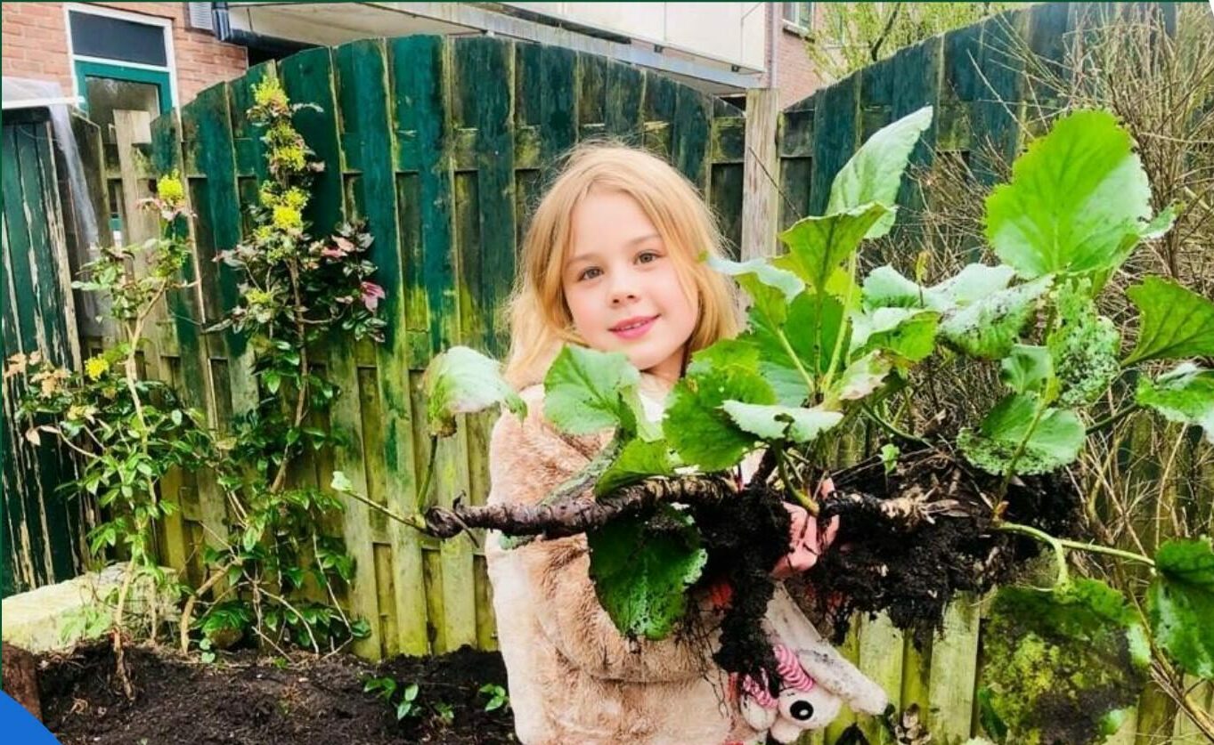 Meisje in tuin houdt grote groene plant vast; houten schutting op de achtergrond.