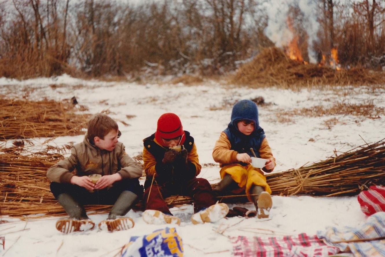 Drie kinderen zitten in de sneeuw bij een vuur, eten en drinken warme dranken.