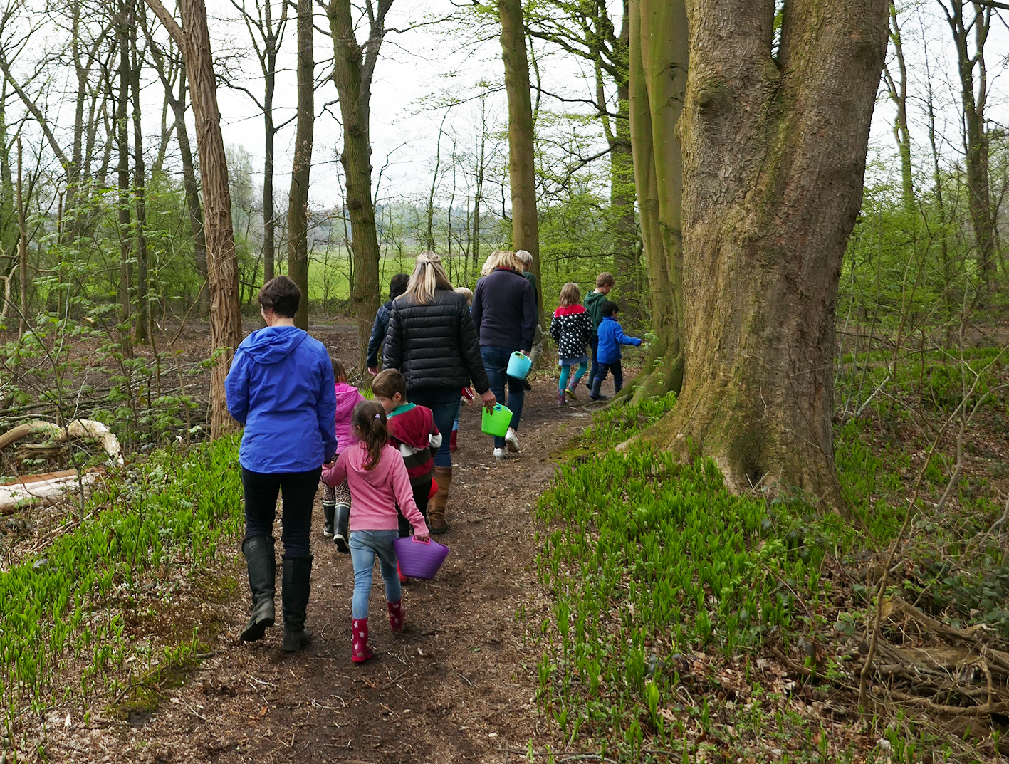 Een groep kinderen en volwassenen wandelt met emmers door een bos.