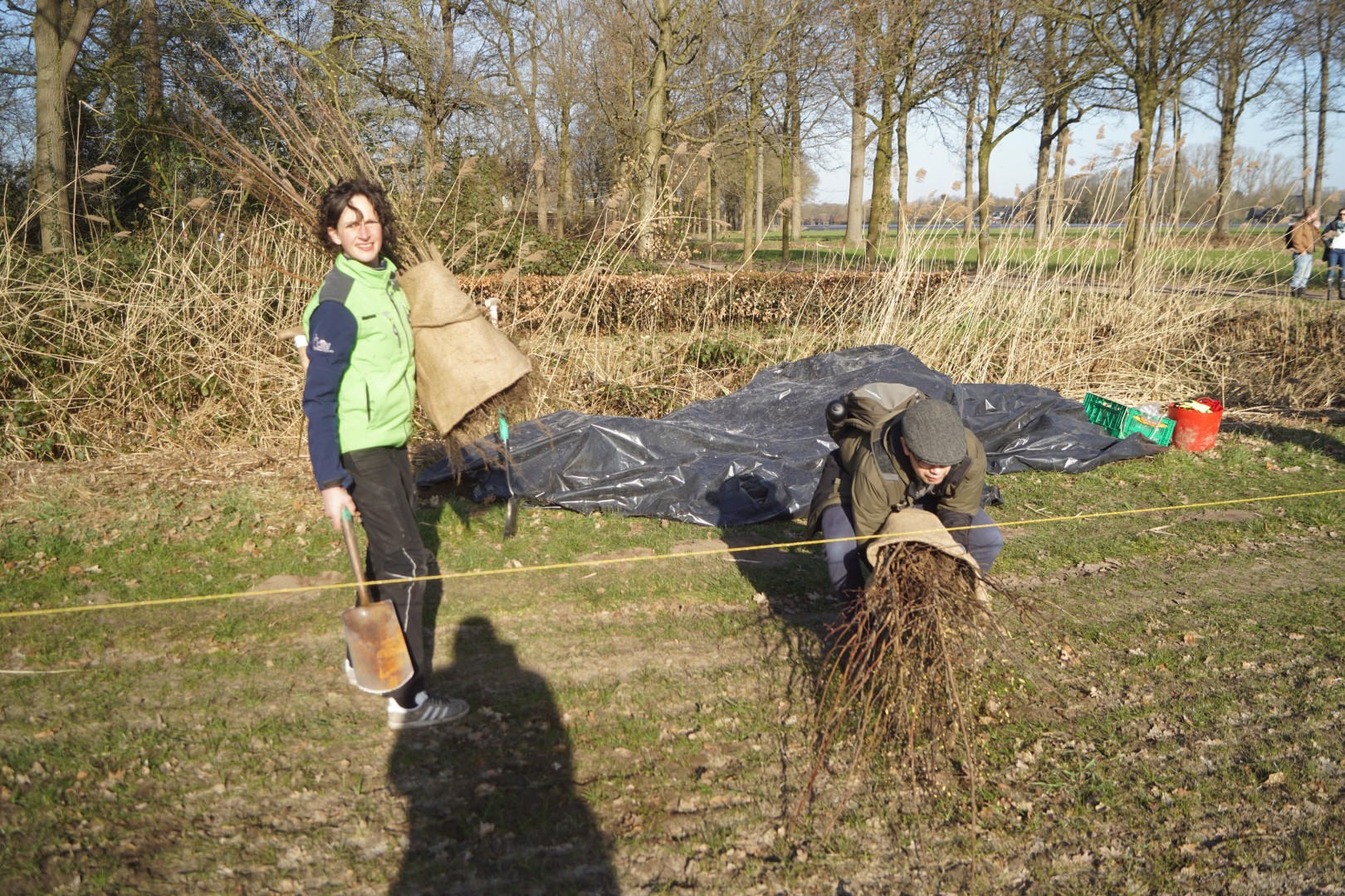 Twee mensen planten bomen in een bosrijk gebied, één draagt een schop en wortels.