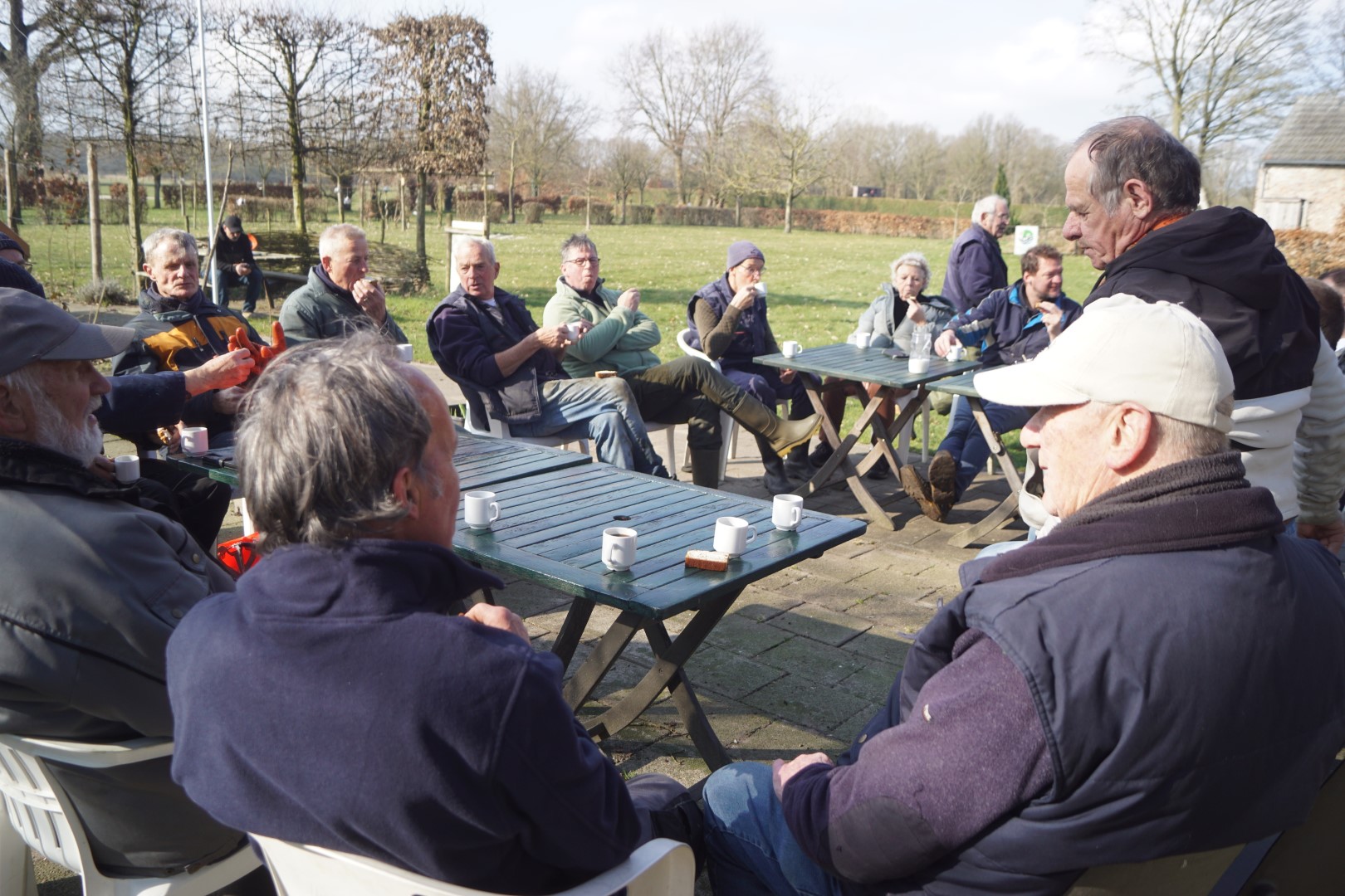 Groep mensen zit buiten rond tafels met koffiekopjes, omgeven door een parklandschap.