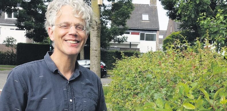 Een lachende persoon met grijs krullend haar staat naast groen struikgewas in een woonwijk.