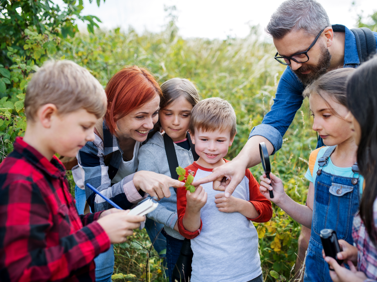 Kinderen en volwassenen onderzoeken een blad met vergrootglazen in een groene buitenomgeving.