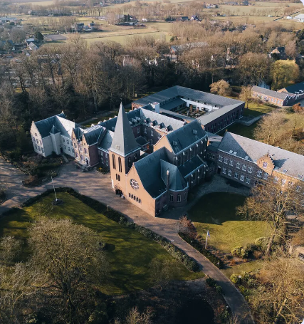 Luchtfoto van groot gebouw met torenspits omgeven door bomen en paden in landelijk gebied.