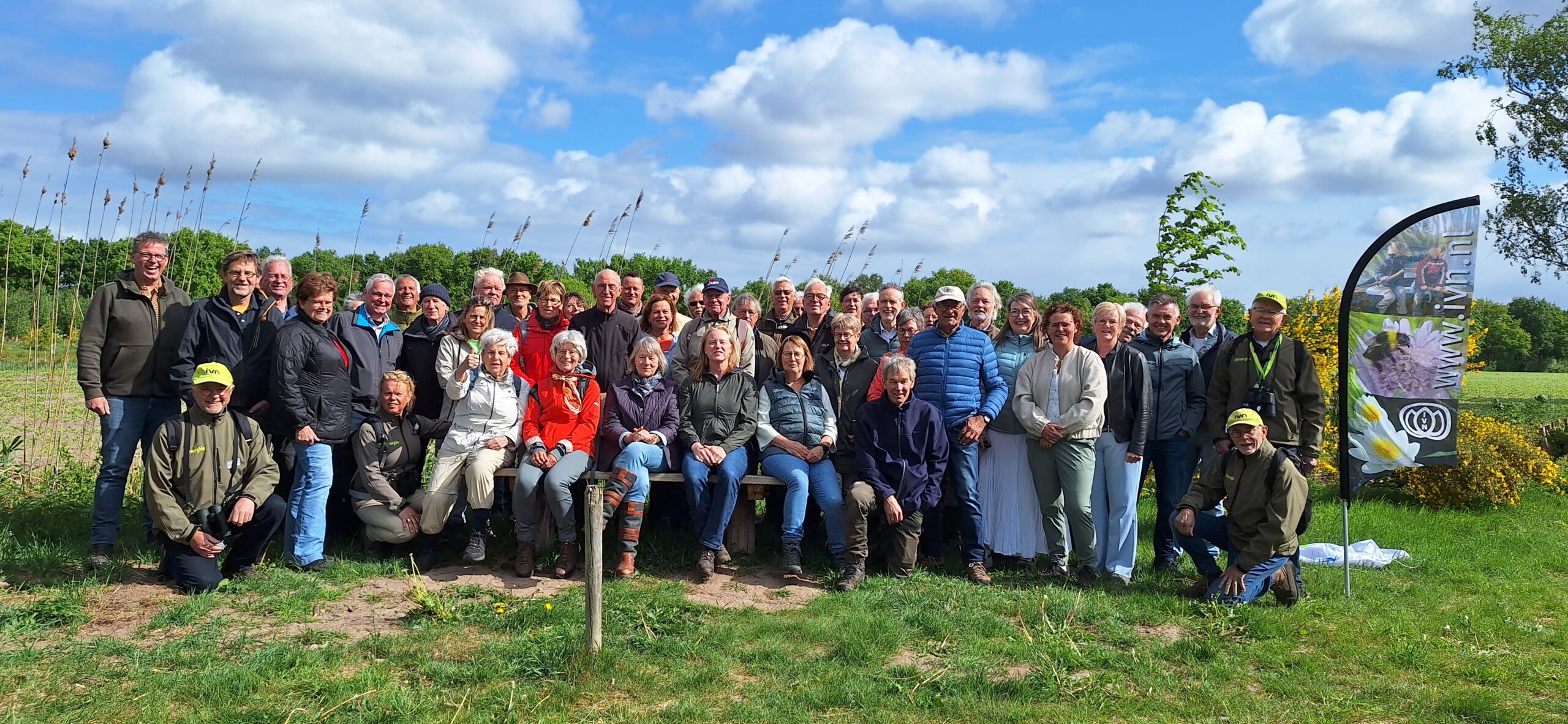 Groepsfoto van mensen buiten in een veld, met een IVN-vlag en een blauwe lucht op de achtergrond.