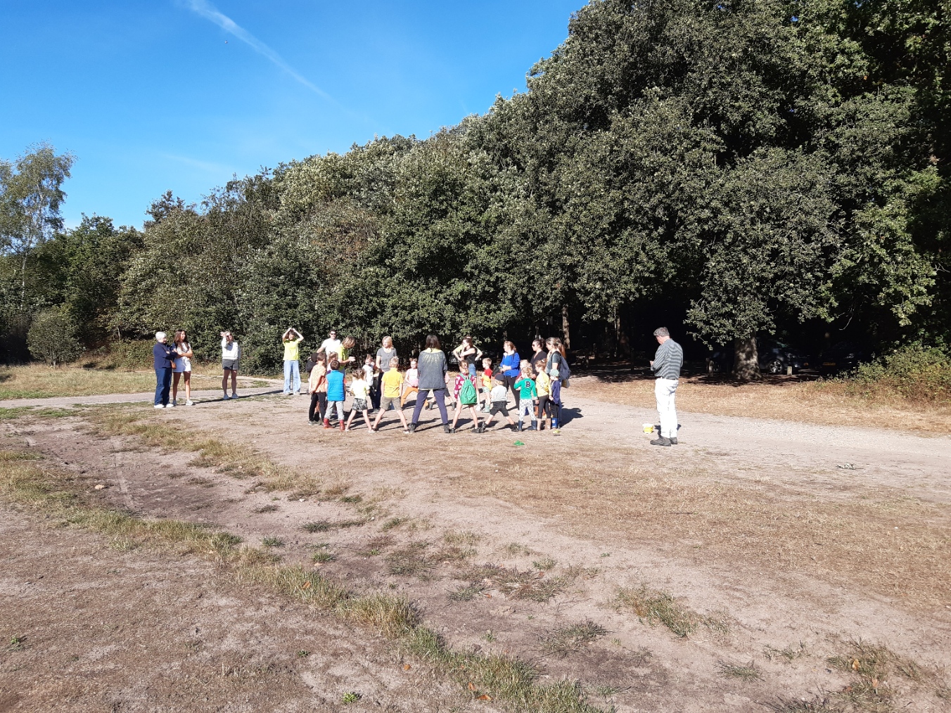 Groep kinderen speelt in een open veld, omringd door volwassenen en bomen onder een blauwe hemel.