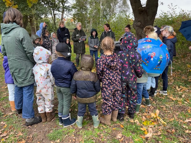 Groep kinderen en volwassenen in regenjassen, verzameld in een kring in een bosrijke omgeving.