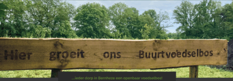 Houten bord met tekst "Hier groeit ons Buurtvoedselbos", met bomen op de achtergrond.