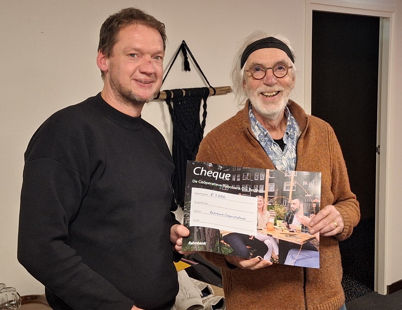 Twee mensen poseren met een cheque van €1000 in een binnenruimte.