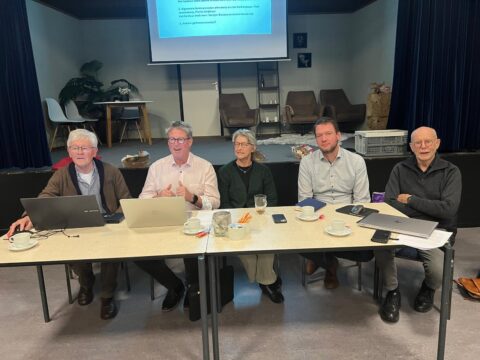 Vijf mensen zitten aan een tafel met laptops en papieren tijdens een presentatie in een vergaderruimte.