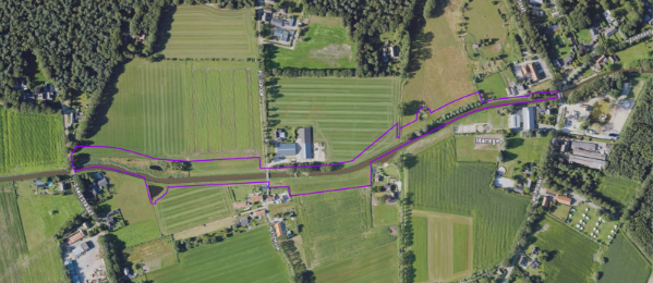 Luchtfoto van groene akkers doorkruist door een paarse lijn; omliggende bebouwing en bos.