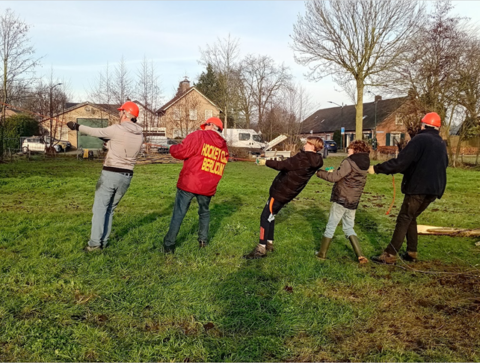 Groep mensen met helmen trekt aan touw op grasveld bij huizen.