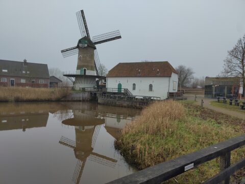 Oude windmolen naast wit gebouw aan rustige vijver, met spiegeling in het water.
