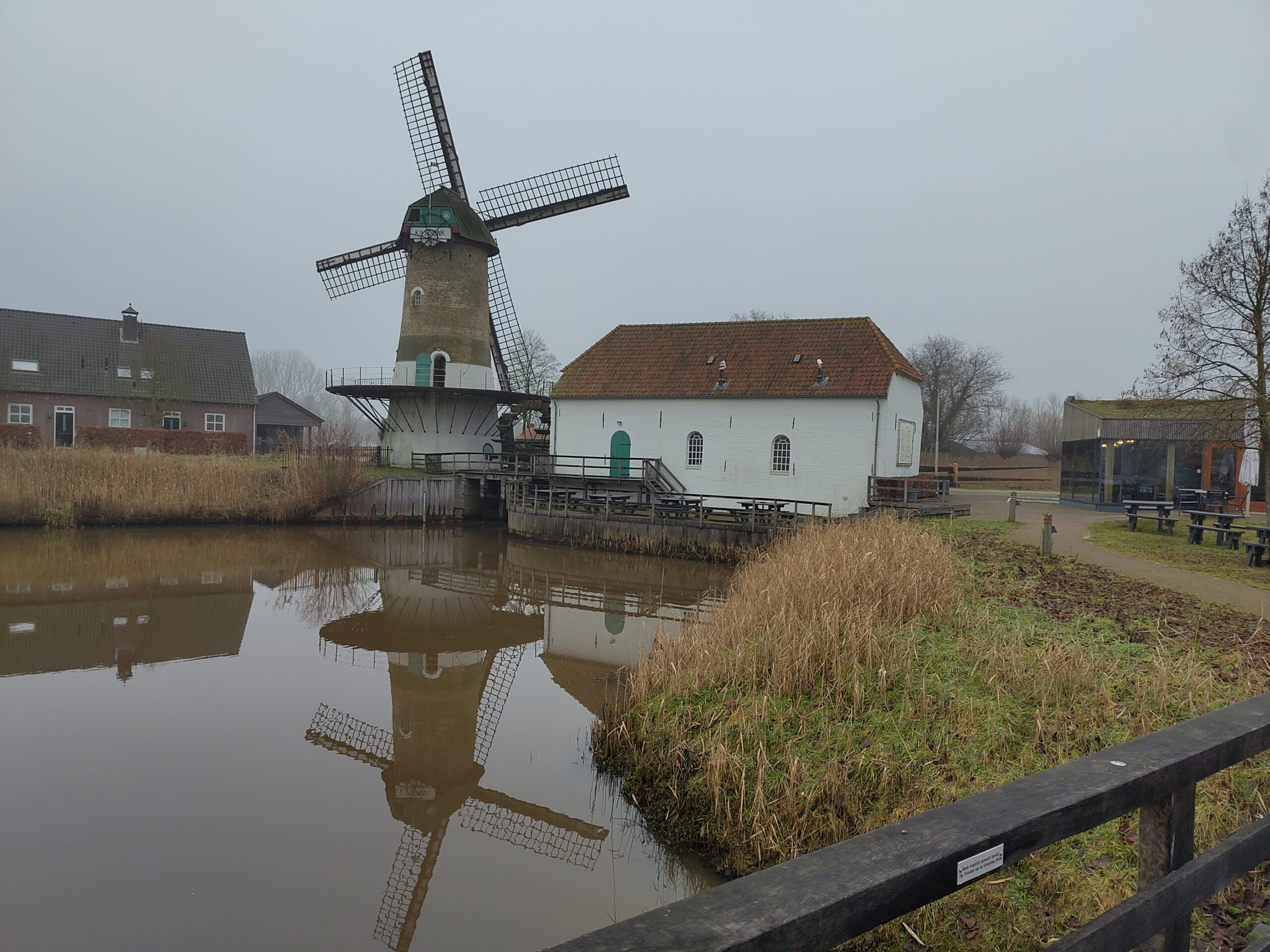 Oude windmolen naast wit gebouw aan rustige vijver, met spiegeling in het water.