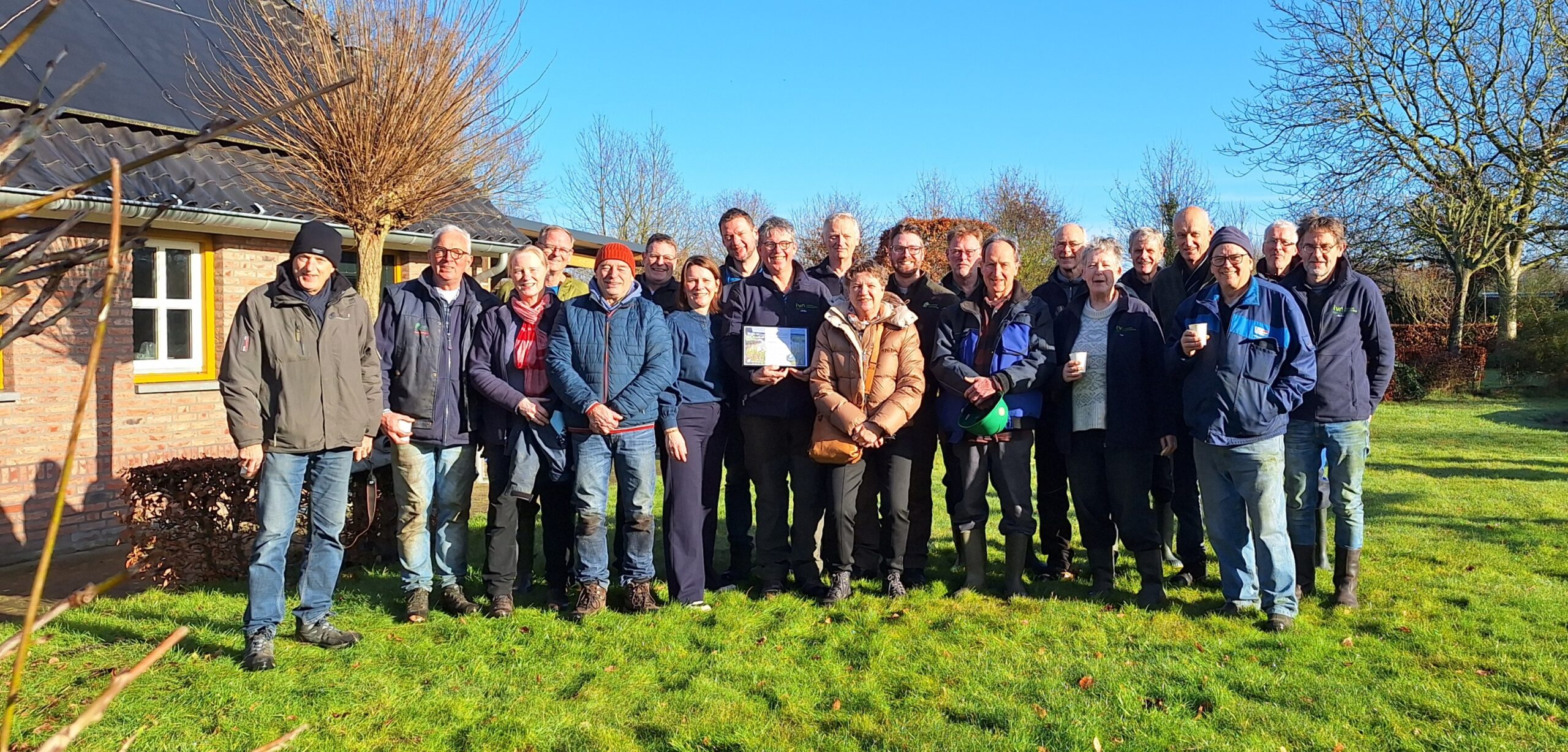 Groep mensen buiten voor een bakstenen huis, zonnige dag, winterkleding.