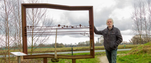Man bij sculptuur van een metalen frame met silhouetten van een landschap, donkere wolken op de achtergrond.