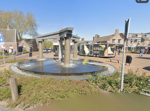 Rond fonteinplein met moderne zuilenstructuur; "KIP&GO" foodtruck op achtergrond.