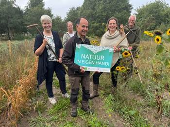 Groep mensen in een veld met zonnebloemen, houden een bord met “Natuur in eigen hand!”.