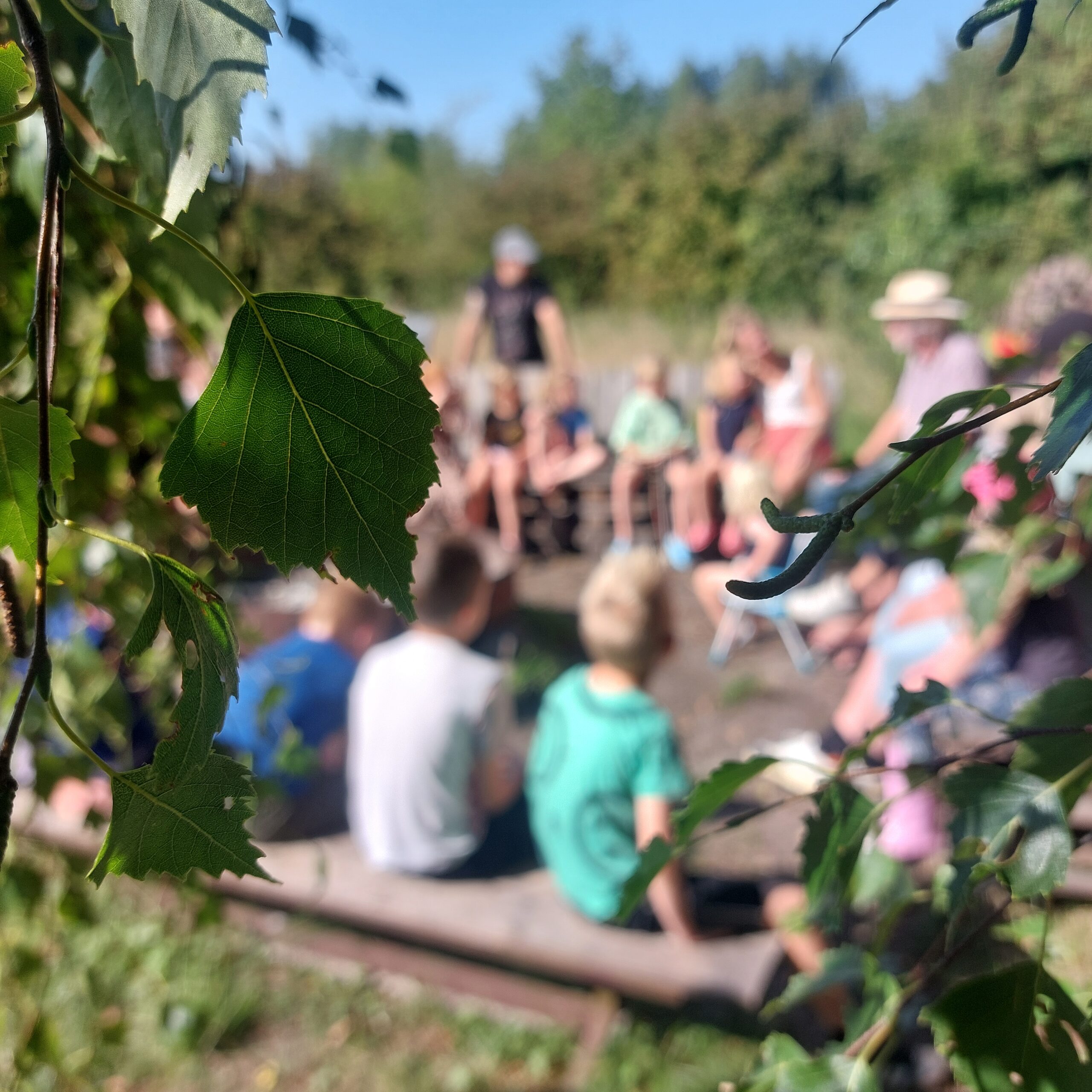 Groep kinderen zit in cirkel buiten, gefotografeerd door bladeren heen.