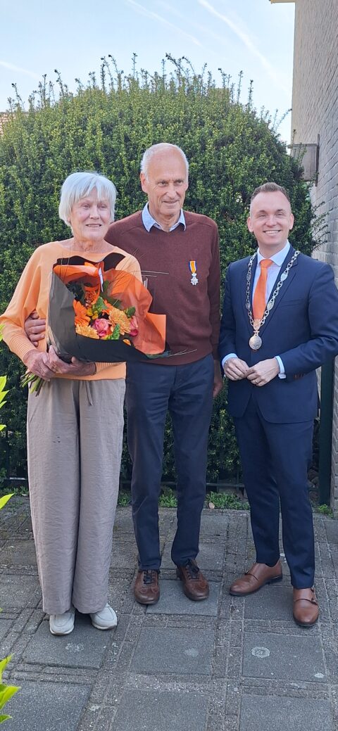 Ouder echtpaar poseert met burgemeester; vrouw met bloemen, man met medaille.
