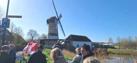 Groep mensen bij een windmolen op een zonnige dag, met parkeerborden en bomen op de achtergrond.