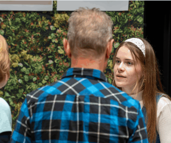 Jonge vrouw in gesprek met man in blauw geruite blouse voor plantenachtergrond.