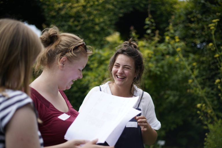 Drie vrolijke vrouwen buiten met papieren in de hand, omgeven door groene struiken.