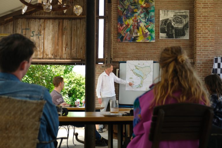 Man presenteert landkaart aan groep zittende mensen in een kunstzinnige, rustieke ruimte.