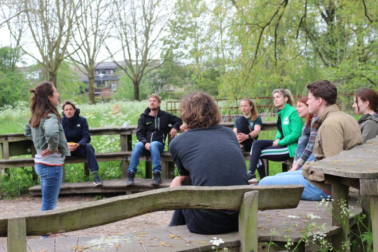 Groep mensen zit buiten in een kring op houten banken terwijl één persoon iets vertelt.