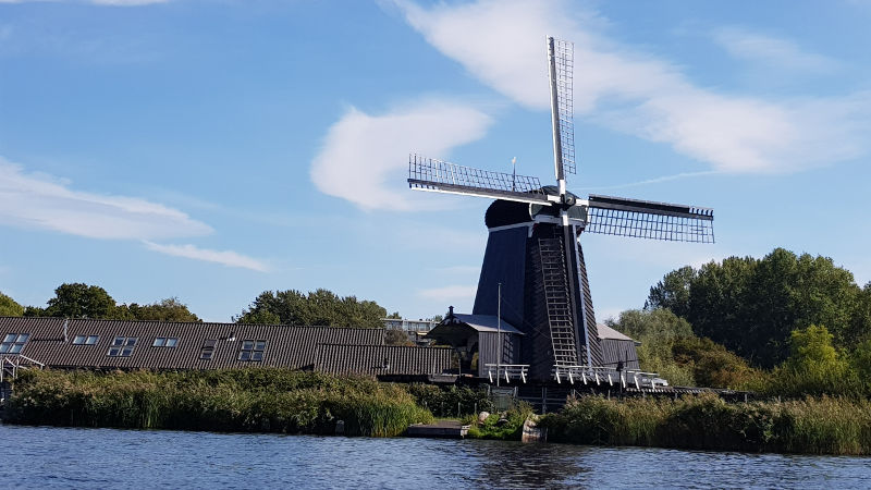Traditionele Nederlandse windmolen aan het water met een heldere blauwe lucht en groene begroeiing.