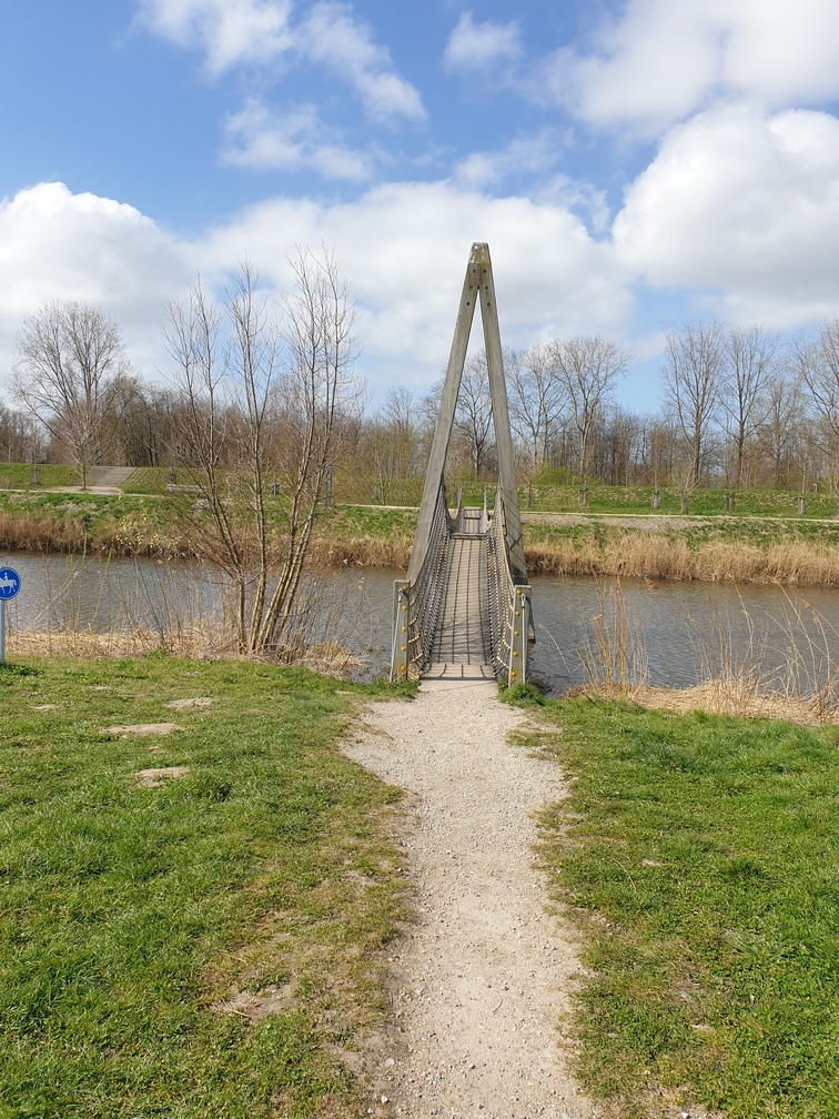 Smalle hangbrug over een rivier, omringd door groen en kale bomen, onder een deels bewolkte hemel.