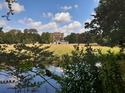 Een statig gebouw achter een grasveld, omringd door bomen, vanaf een vijver gezien onder een blauwe lucht.