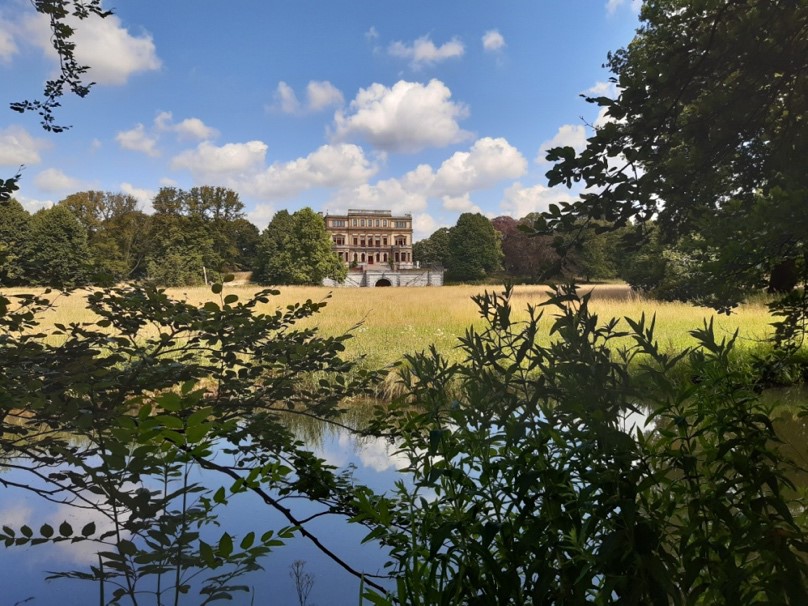 Een statig gebouw achter een grasveld, omringd door bomen, vanaf een vijver gezien onder een blauwe lucht.