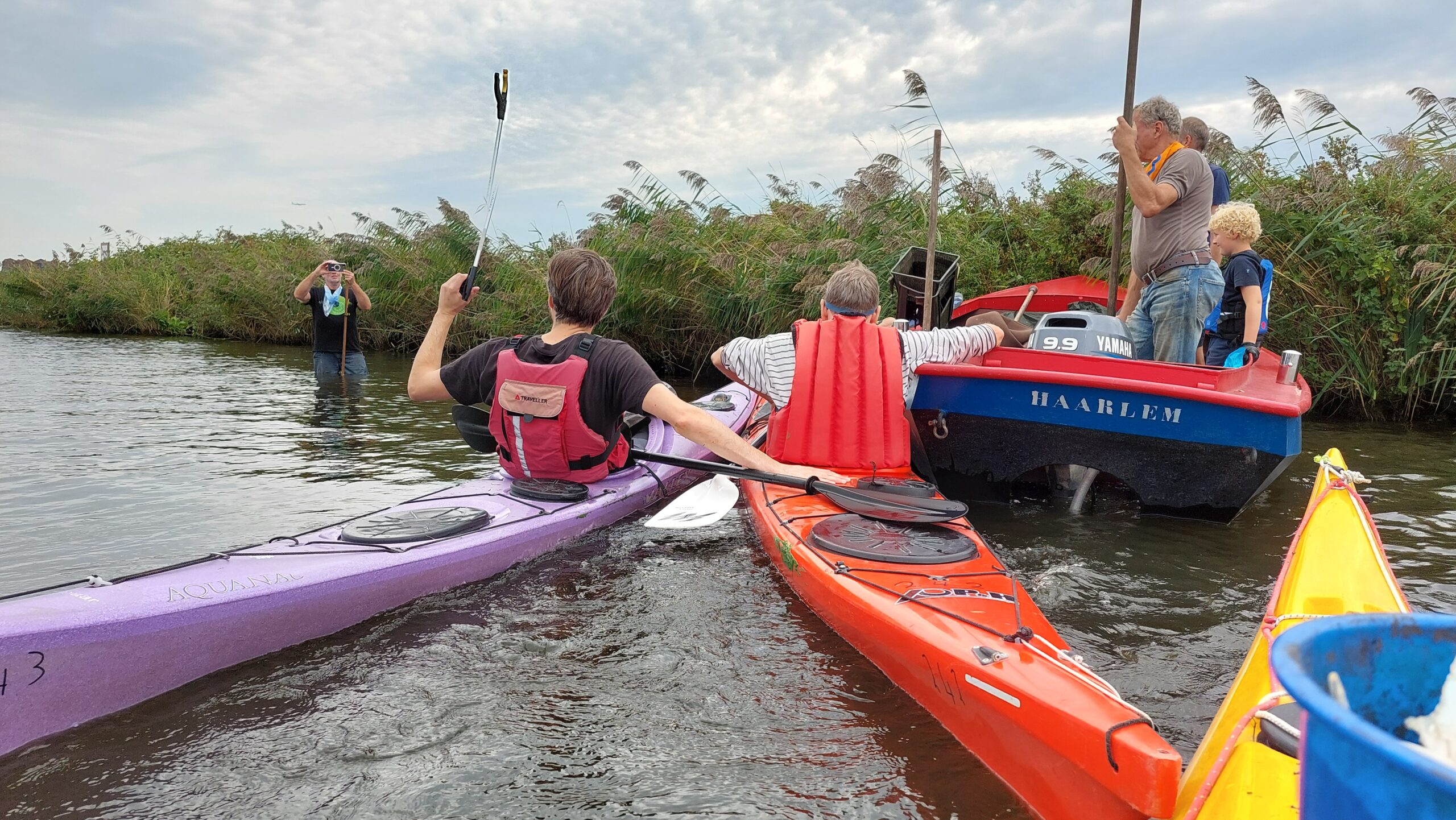 Twee mensen in kajaks bij een boot op een rivier; iemand maakt een foto vanaf de oever.
