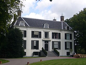 Groots, wit historisch landhuis met donkere luiken en een trap, omringd door groen en wolkenlucht.