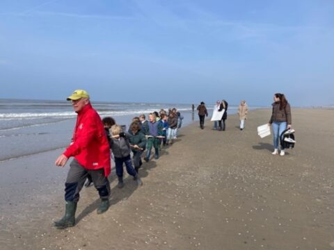 Een groep kinderen volgt een man in een rode jas langs de kustlijn van een strand.