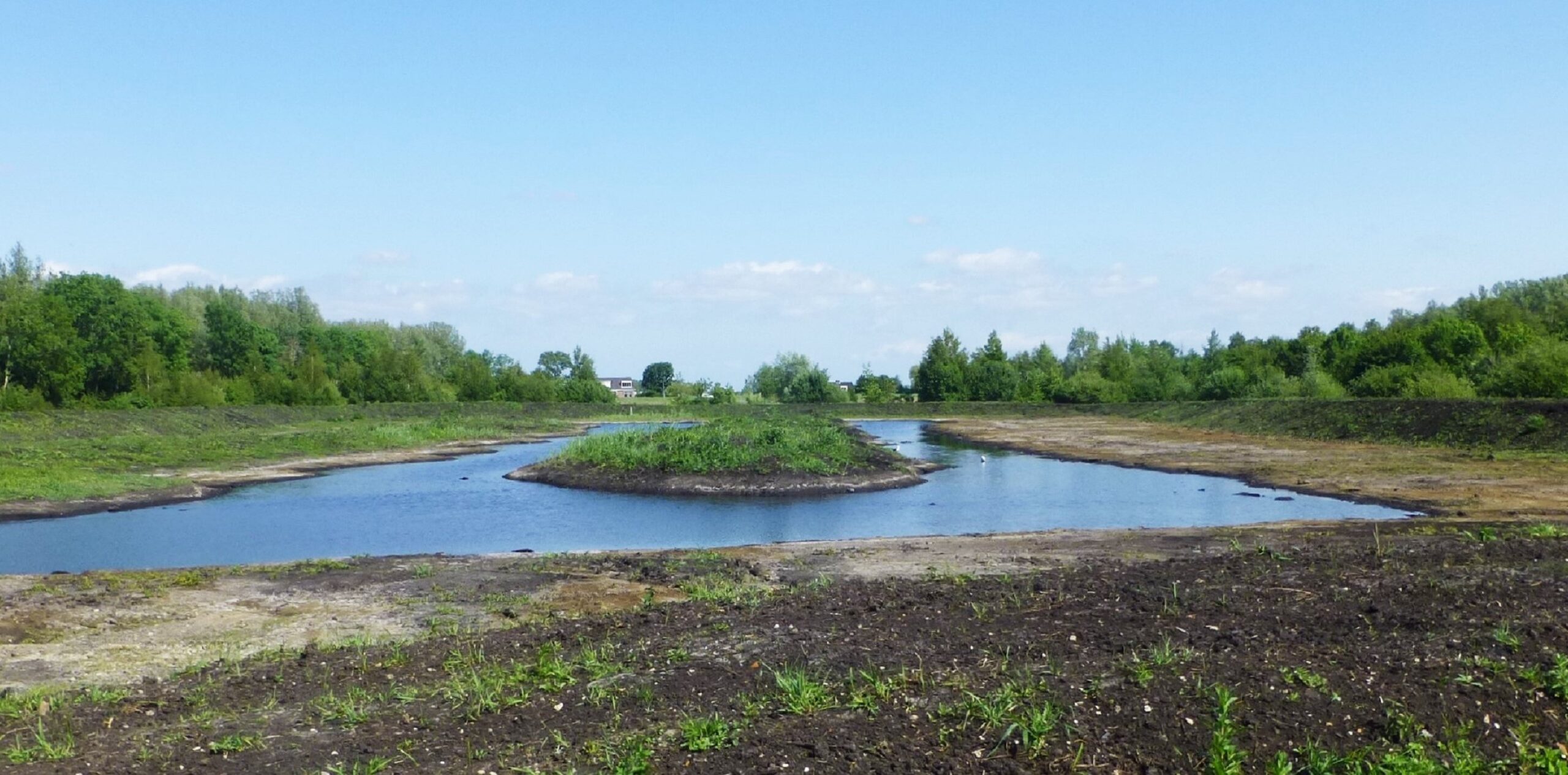 Een vijver met een eiland, omgeven door groen landschap onder een heldere blauwe lucht.