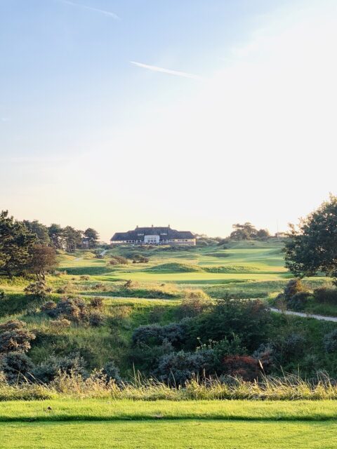 Uitzicht op een uitgestrekt, groen golfterrein met clubhuis en bomen onder blauwe hemel.